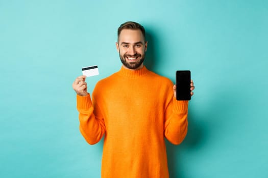 Online shopping. Happy attractive guy showing mobile phone screen and credit card, smiling satisfied, standing over light blue background.