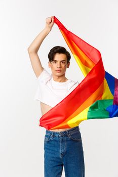 Vertical view of happy queer person in crop top and jeans waving raised rainbow flag, celebrating lgbtq holiday, standing over white background.