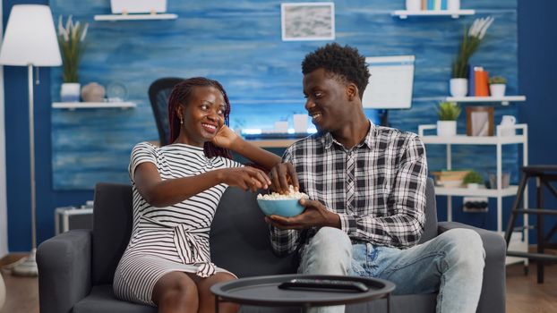 POV of black people having fun while watching TV in living room. African american couple laughing at comedy looking at camera. Cheerful lovers of african ethnicity eating popcorn