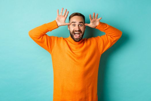 Image of handsome caucasian man making funny faces, mocking someone and smiling, standing against light blue background.