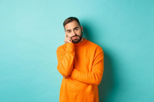Sad and gloomy man with beard, looking indesicive and sad, thinking about something and sulking, standing over turquoise background.