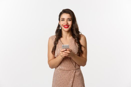 Christmas party and celebration concept. Happy young woman in elegant dress using mobile phone and smiling, shopping online, standing over white background.