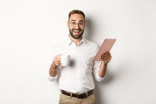 Excited manager reading on digital tablet, working and drinking coffee, standing against white background.