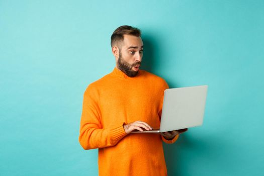 Freelance and technology concept. Image of handsome adult man staring at laptop with amazement, shopping online or working remote, standing over turquoise background.