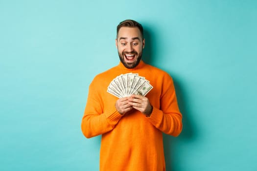 Image of man looking happy at money, smiling amazed, standing over light blue background.