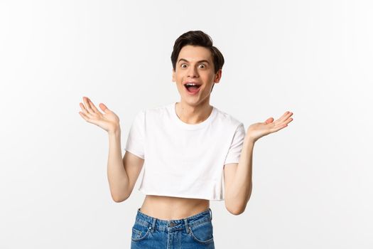 Lgbtq and pride concept. Handsome young man in crop top raising hands up and looking surprised, rejoicing of good news, standing over white background.