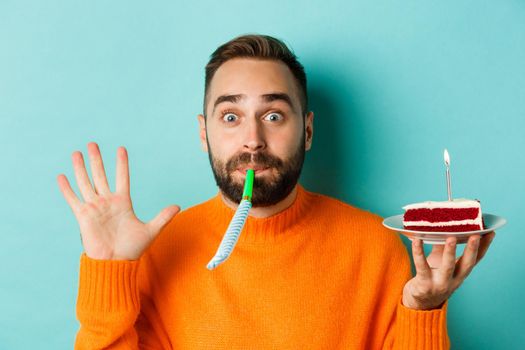 Close-up of funny adult man celebrating his birthday, holding bday cake with candle, blowing party wistle and rejoicing, standing over light blue background.