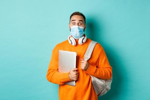 Handsome caucasian man with headphones and backpack, holding laptop and wearing medical mask, looking surprised, standing over light blue background.