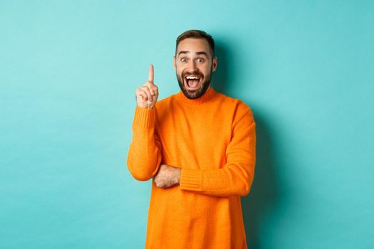 Handsome caucasian man having an idea, raising finger up and saying his plan, standing excited against turquoise background.