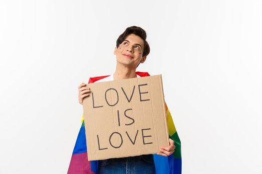 Dreamy young queer person smiling and looking at upper left corner, holding love is love sign for pride parade, wearing Rainbow flag, white background.