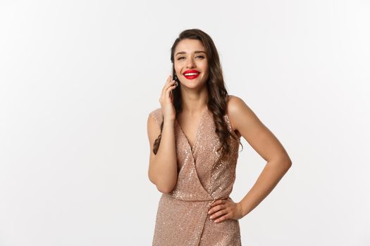 Christmas party and celebration concept. Attractive young woman with red lips, wearing luxury dress, talking on mobile phone and smiling, standing over white background.