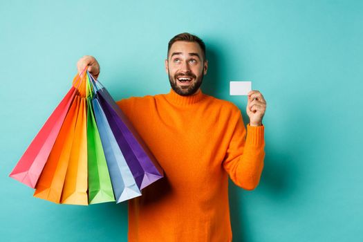 Happy aduly man showing credit card and shopping bags, standing against turquoise background.