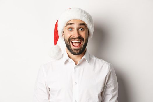 Close-up of happy bearded guy in santa hat, looking surprised, celebrating christmas, standing over white background.