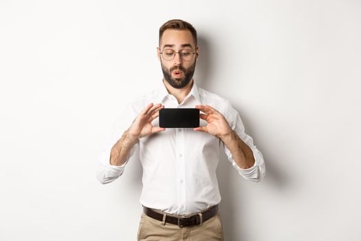 Amused handsome guy showing mobile screen, looking excited at online website, standing over white background.