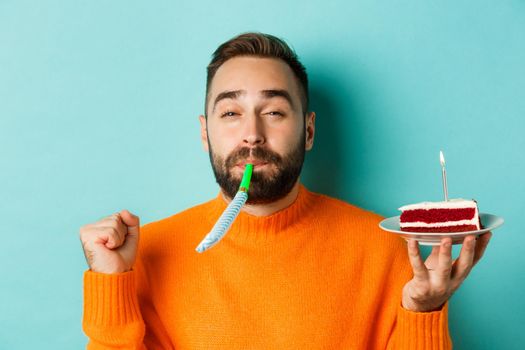 Close-up of funny adult man celebrating his birthday, holding bday cake with candle, blowing party wistle and rejoicing, standing over light blue background.