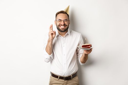 Holidays and celebration. Happy man making wish on birthday cake, cross fingers and smiling excited, having b-day party, white background.