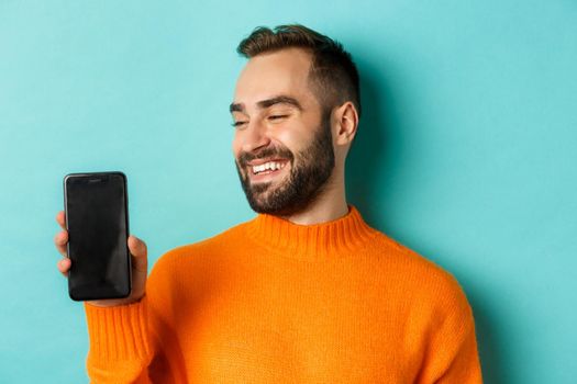 Close-up of young bearded man showing phone screen and looking satisfied, wearing orange sweater, standing against studio background.