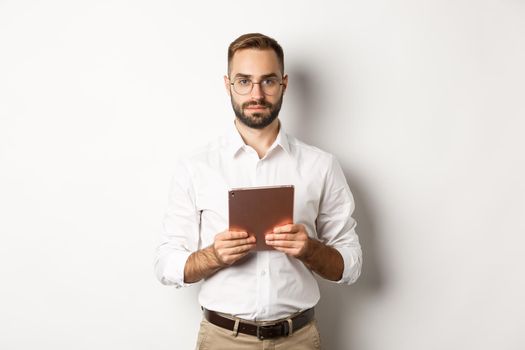 Serious employer working with digital tablet, reading in glasses, standing over white background.