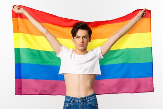 Out and proud. Happy and confident gay man with glitter on face raising lgbtq rainbow flag and smiling at camera, standing in crop top, white background.