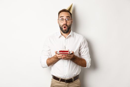 Holidays and celebration. Happy man having birthday party, making wish on b-day cake and smiling, standing against white background.