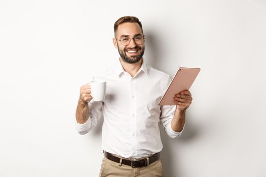 Satisfied boss drinking tea and using digital tablet, reading or working, standing over white background.