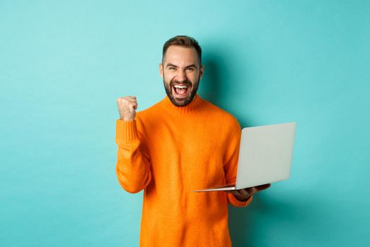 Freelance and technology concept. Lucky man winner celebrating, winning online, showing fist pump and holding laptop, standing over light blue background.