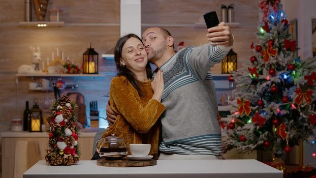 Cheerful couple taking pictures on christmas eve with smartphone. Married man and woman making memories with selfies for holiday season celebration. People in festive decorated kitchen