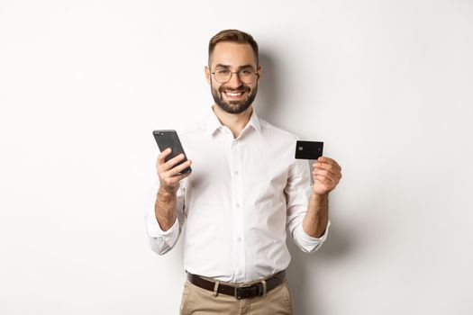 Business and online payment. Smiling male entrepreneur shopping with credit card and mobile phone, standing over white background.
