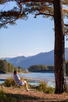 A woman is sitting on a folding chair on the bank of a mountain river on a nice, warm day under a large tree. A calm and quiet place to relax and reflect. Equipment and a tourist's rest.