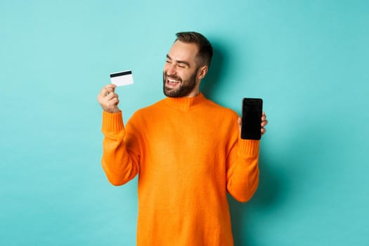 Online shopping. Satisfied man using credit card and showing mobile screen, looking pleased, standing over light blue background.