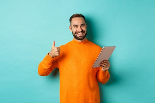 Satisfied adult man smiling, using digital tablet and showing thumb-up, approve and agree, standing against turquoise background.