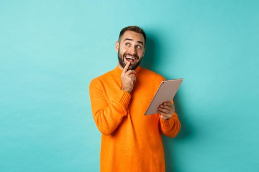 Handsome man thinking and using digital tablet, shopping online and pondering ideas, looking at upper left corner, standing over light blue background.