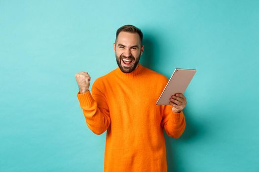 Cheerful winning man holding digital tablet, rejoicing and celebrating victory in game, making fist pump gesture, standing over light blue background.