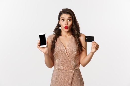 Online shopping and holidays concept. Excited beautiful lady in elegant dress showing mobile screen and credit card, standing over white background.