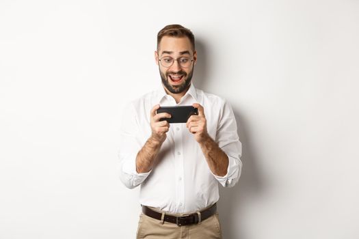 Man looking excited and surprised at mobile phone, holding smartphone horizontally, standing over white background.