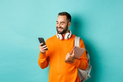 Handsome man student with headphones and backpack, holding digital tablet, reading message on mobile phone, standing against light blue background.