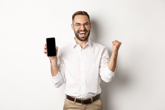 Successful business man showing mobile screen, rejoicing on winning online prize, achieve app goal, standing against white background.