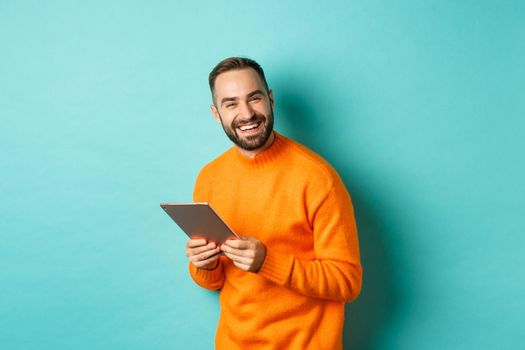 Handsome bearded man using digital tablet, laughing at camera, standing happy against light blue background.