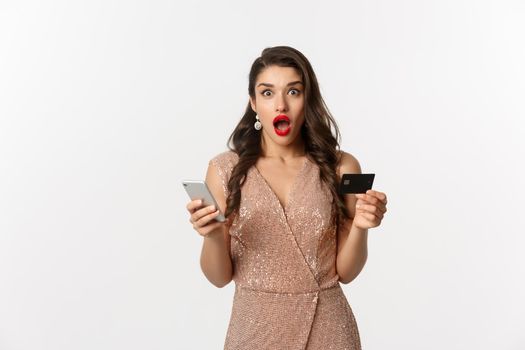 Online shopping and holidays concept. Surprised woman in glamour dress, buying in internet with credit card and mobile phone, looking amazed, white background.