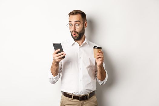 Businessman drinking coffee and looking surprised at message on mobile phone, standing amazed over white background.