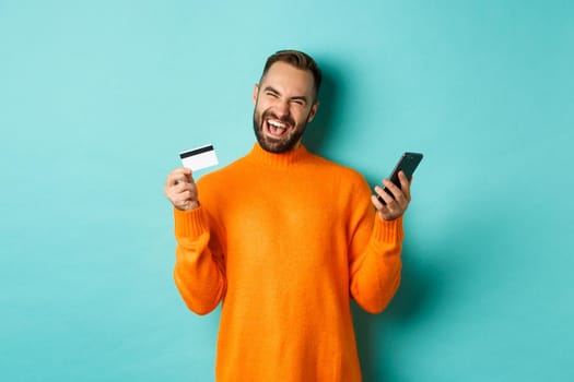 Online shopping. Happy young man using mobile phone and credit card, paying internet, light blue background.