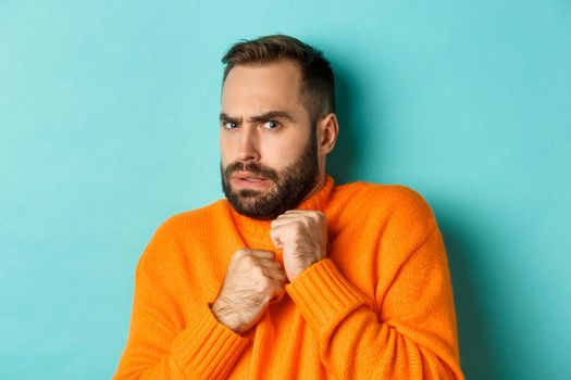 Close-up of scared bearded man looking startled and displeased, jumping from fear, standing over light blue background.