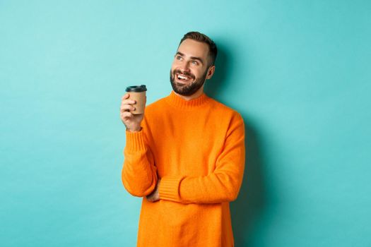 Thoughtful smiling man looking at upper left corner, having idea while drinking coffee from takeaway cup, standing in orange sweater against turquoise background.