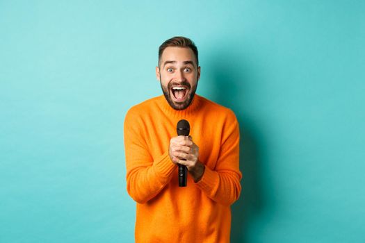 Handsome adult man perform song, singing into microphone, standing against turquoise background.