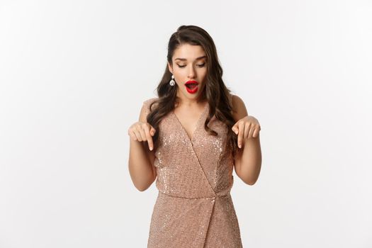 Amazed glamour girl in party dress, looking and pointing down amazed, showing christmas advertisement, standing over white background.