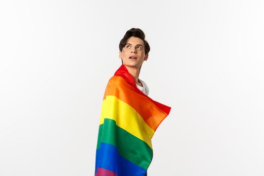 Human rights and lgbtq community concept. Beautiful young androgynous man with glitter on face, wearing rainbow pride flag and looking at upper left corner, white background.