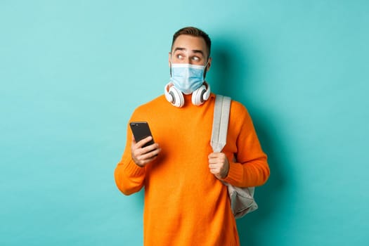 Young man in face mask using mobile phone, holding backpack, staring left amazed, standing against light blue background.