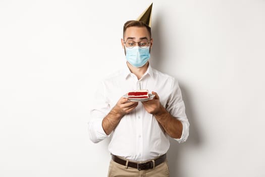 Covid-19, social distancing and celebration. Man looking excited at birthday cake, wearing medical mask from coronavirus pandemic, white background.