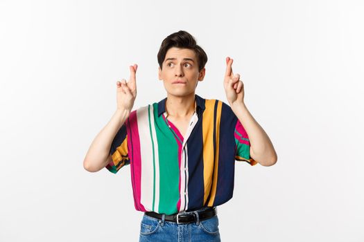 Hopeful male model looking worried at upper left corner, cross fingers for good luck, making wish, standing over white background.
