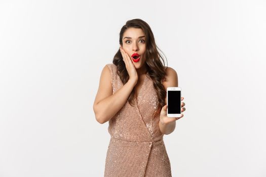 Online shopping. Elegant woman in luxury dress showing smartphone screen, looking amazed at camera, white background.